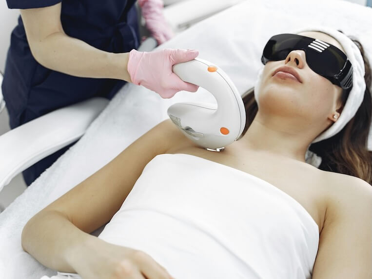 From above female cosmetologist providing cosmetic procedures with laser to female client in salon