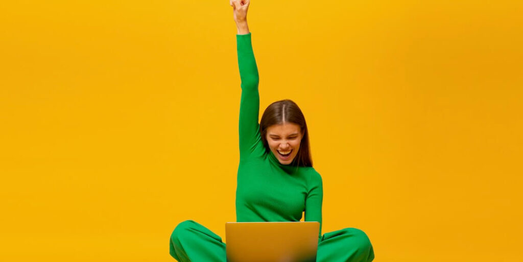 winner excited happy girl sitting on floor with laptop raising one hand in the air if saying yes isolated on yellow background FEATURED IMAGE