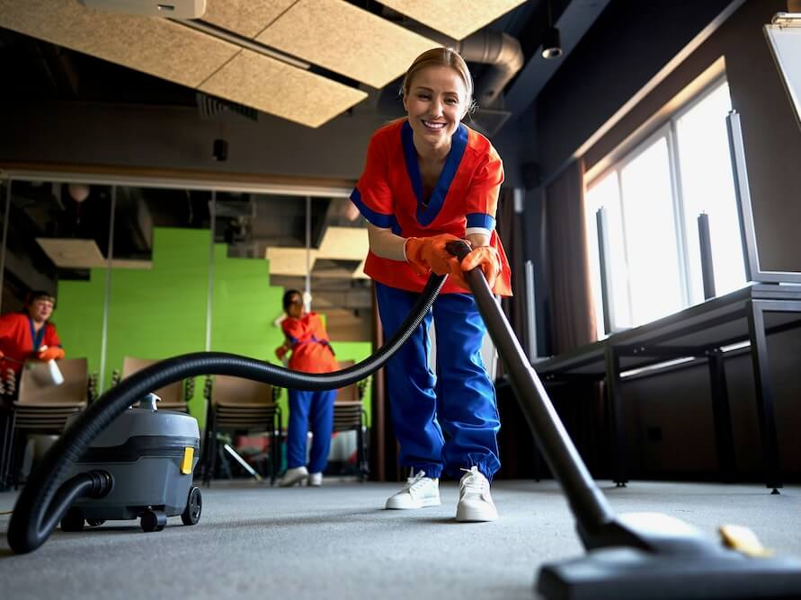 smiling pleased female worker vacuuming the carpet flooring with a corded bagless canister vacuum cleaner