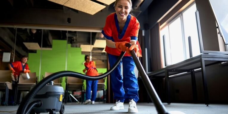smiling pleased female worker vacuuming the carpet flooring with a corded bagless canister vacuum cleaner FEATURED IMAGE