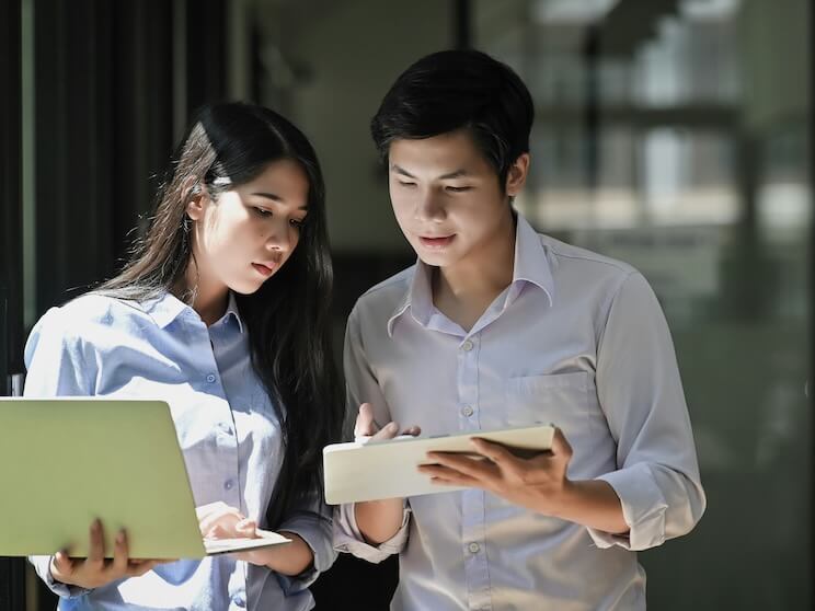 photo of young business startup couple standing together holding and looking at laptop and tablet while talking/discussing or brainstorming about their business 