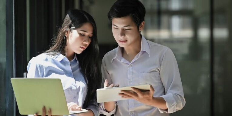photo of young business startup couple standing together holding and looking at laptop and tablet while talking/discussing or brainstorming about their business | FEATURED IMAGE