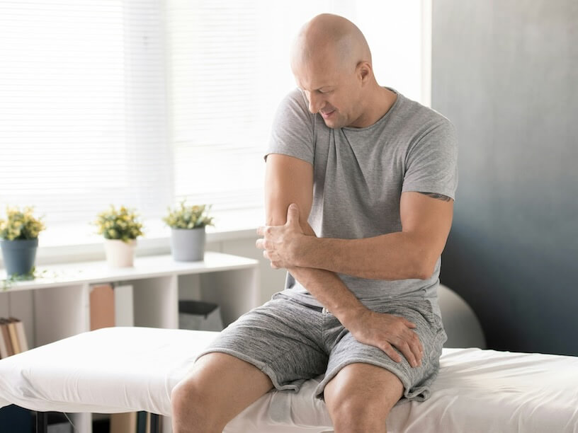 bald man touching painful elbow and trying to massage it before physical exercise while sitting on medical couch in clinic and waiting for doctor