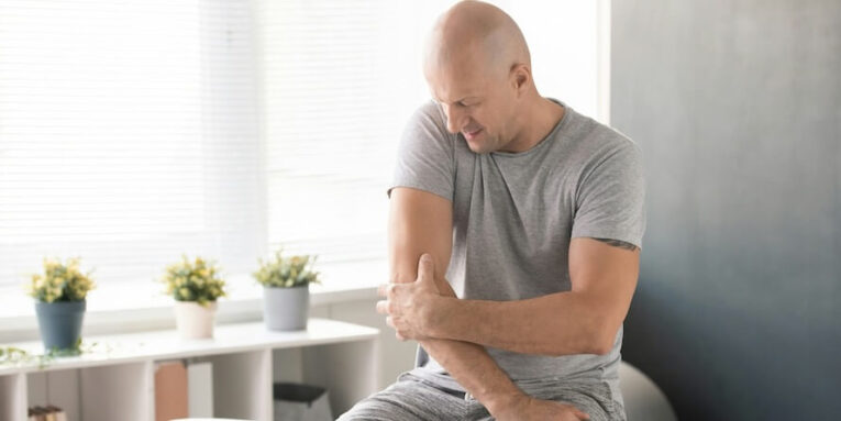 bald man touching painful elbow and trying to massage it before physical exercise while sitting on medical couch in clinic and waiting for doctor FEATURED IMAGE