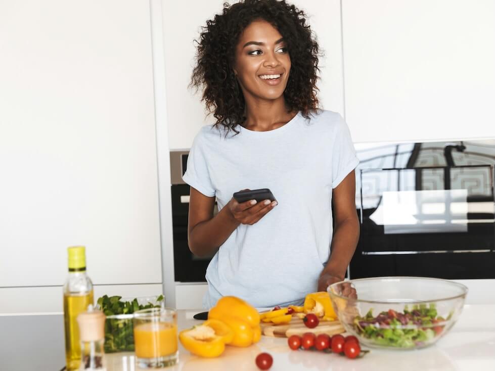 portrait of a smiling African american woman using mobile phone while making a healthy salad in the kitchen