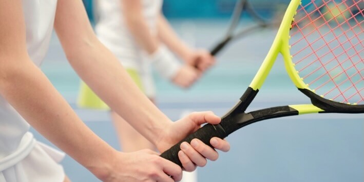 hands of teenage girl in white activewear standing on stadium against field and holding tennis racket ready to push the ball during play FEATURED IMAGE