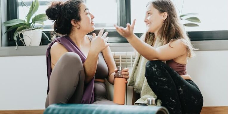 two women sitting on a yoga mat talking to each other FEATURED IMAGE