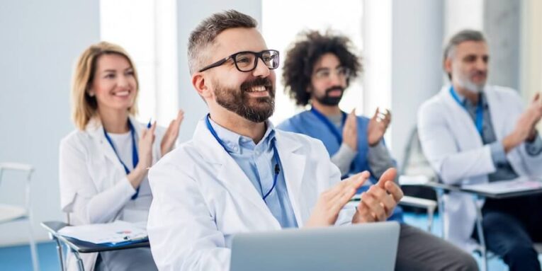 a group of doctors listening to presentation on medical conference clapping FEATURED IMAGE