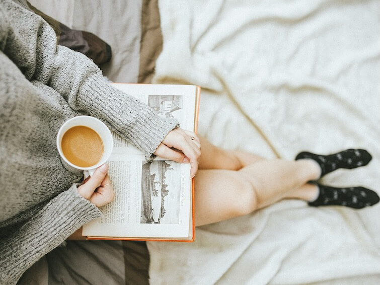 woman holding a cup of coffee at right hand and reading book on her lap while holding It open with her left hand in a well lit room