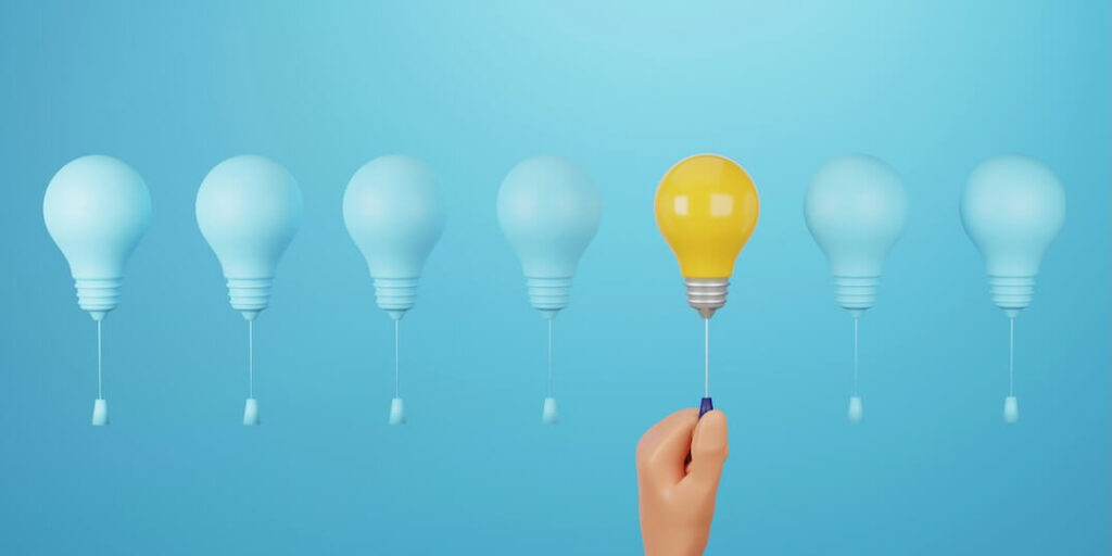 hand turning on a glowing yellow light bulb between blue light bulbs on a blue background different creativity outstanding idea thinking 3d render illustration FEATURED IMAGE