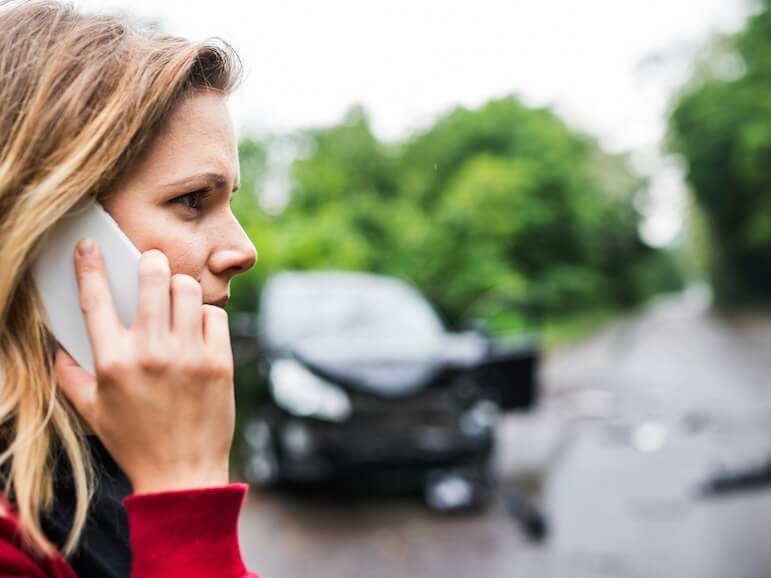 a close up of a young woman on the phone standing by the damaged car after a car accident