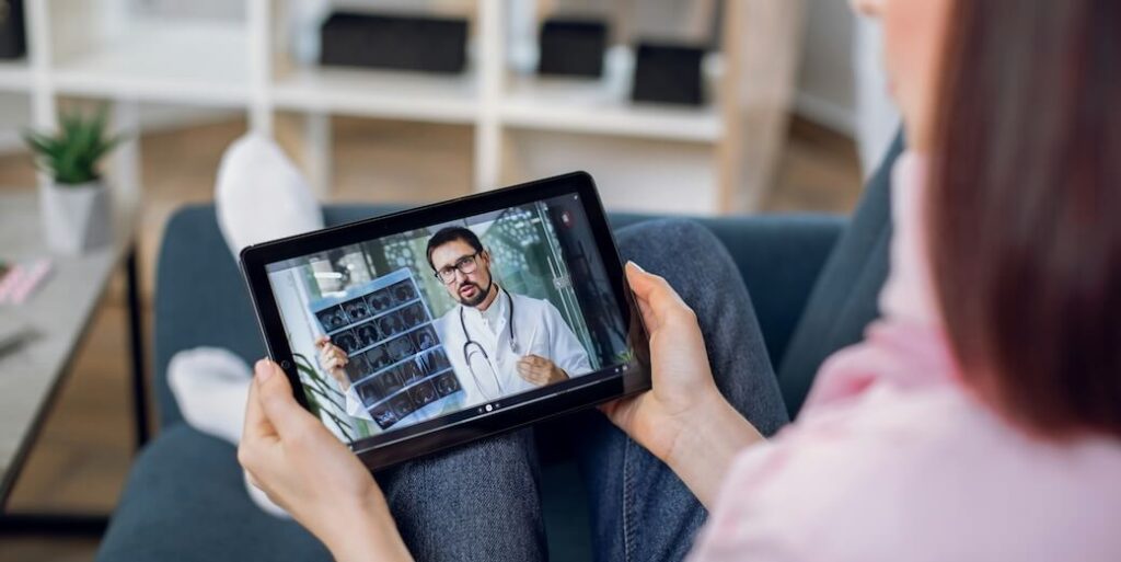 telemedicine remote medicine concept back shoulder view of tablet pc screen with confident male doctor holding xray scan and consulting his patient staying at home woman sits on sofa at home FEATURED IMAGE
