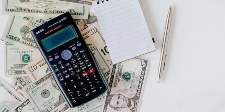 Calculator and Notepad placed over Stack of Paper Bills FEATURED IMAGE