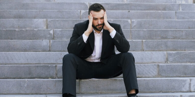 Upset young business man having stress and sitting on stairs in street. Businessman having deal problems at work concept FEATURE IMAGE
