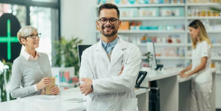 young male pharmacist posing while working in a pharmacy while his female colleague is selling prescription medications to senior female customer FEATURED IMAGE