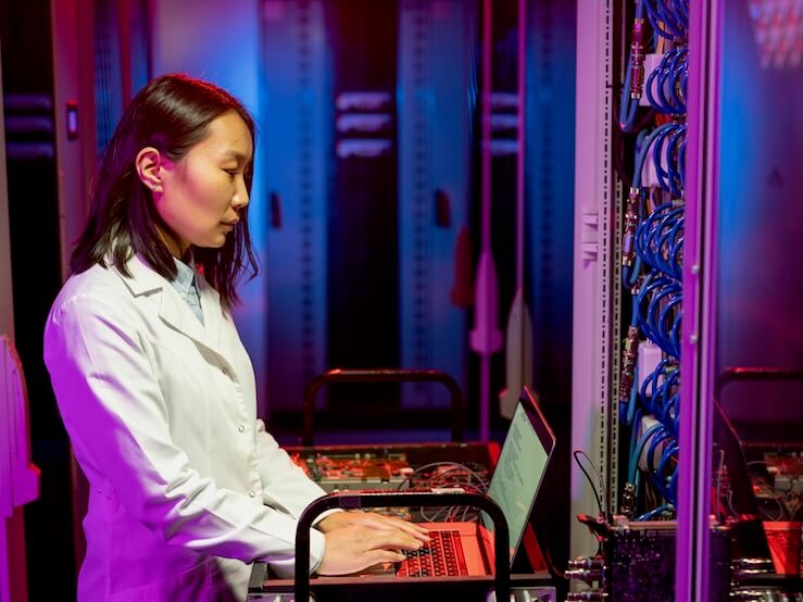 serious young asian it specialist in white coat standing at cart and using laptop while performing diagnostic tests