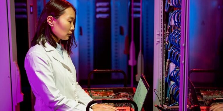 serious young asian it specialist in white coat standing at cart and using laptop while performing diagnostic tests FEATURED IMAGE