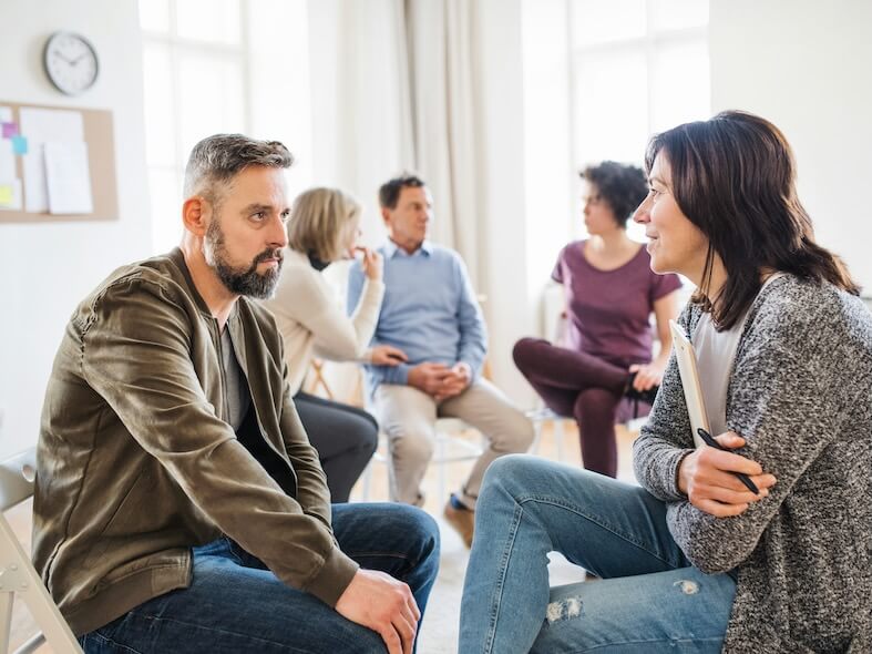 a senior counselor with clipboard talking to a man during group therapy