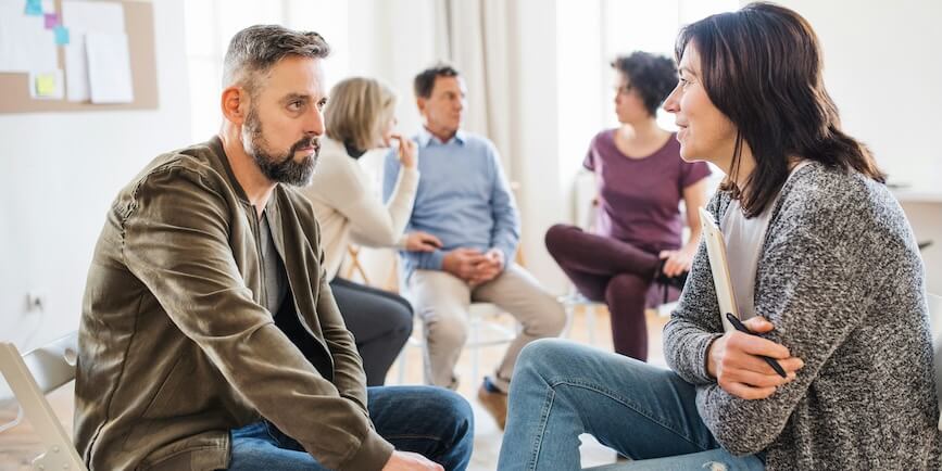 a senior counselor with clipboard talking to a man during group therapy FEATURE IMAGE