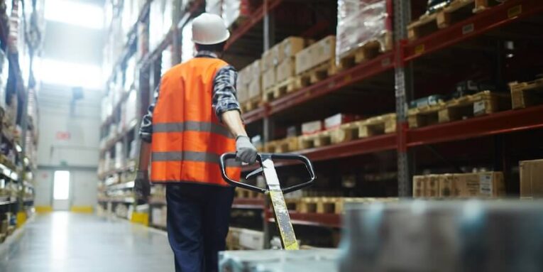worker in helmet and uniform pulling forklift with packed goods FEATURED IMAGE