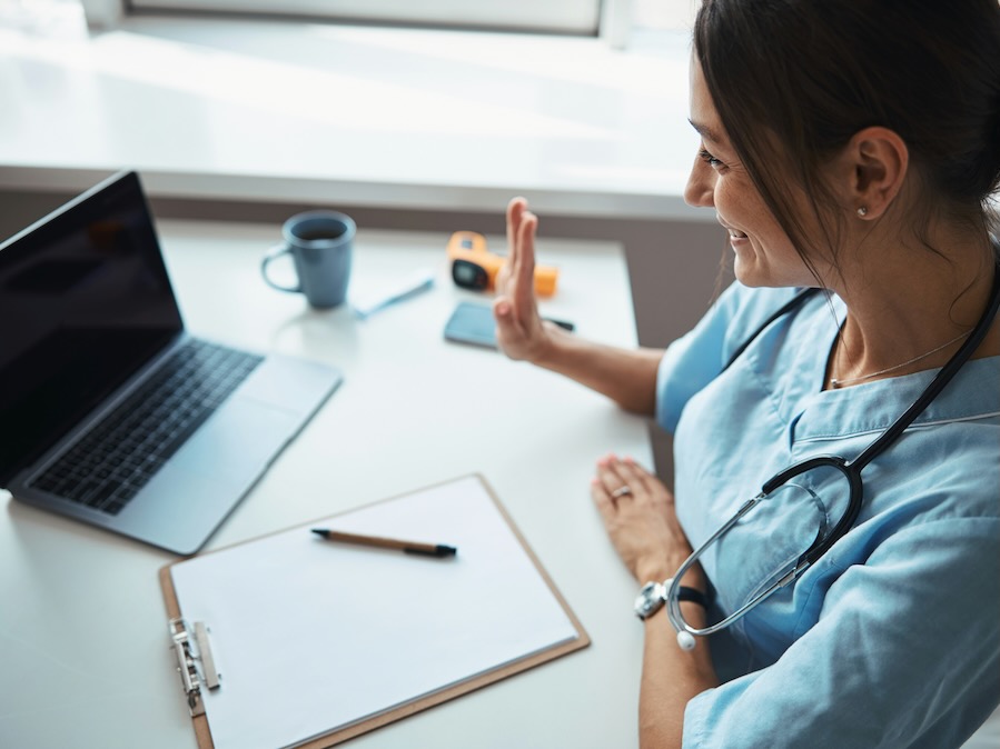 joyful young woman physician doing hello gesture and smiling while sitting at the table with notebook and giving online consultation