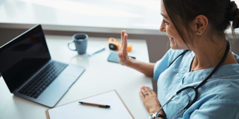 joyful young woman physician doing hello gesture and smiling while sitting at the table with notebook and giving online consultation FEATURED IMAGE