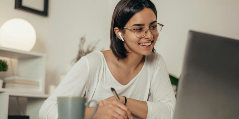 beautiful woman writing in a notebook and using a laptop while managing her business and working remotely from home FEATURED IMAGE
