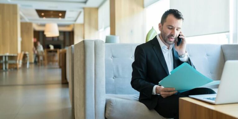 handsome mid adult businessman busy on call while reading business plan from file in hotel lobby FEATURED IMAGE