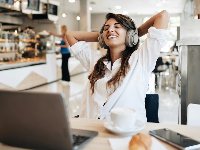 beautiful and happy young woman sitting and eating delicious rolls in bakery or fast food she also using her laptop and listening to music with headphones