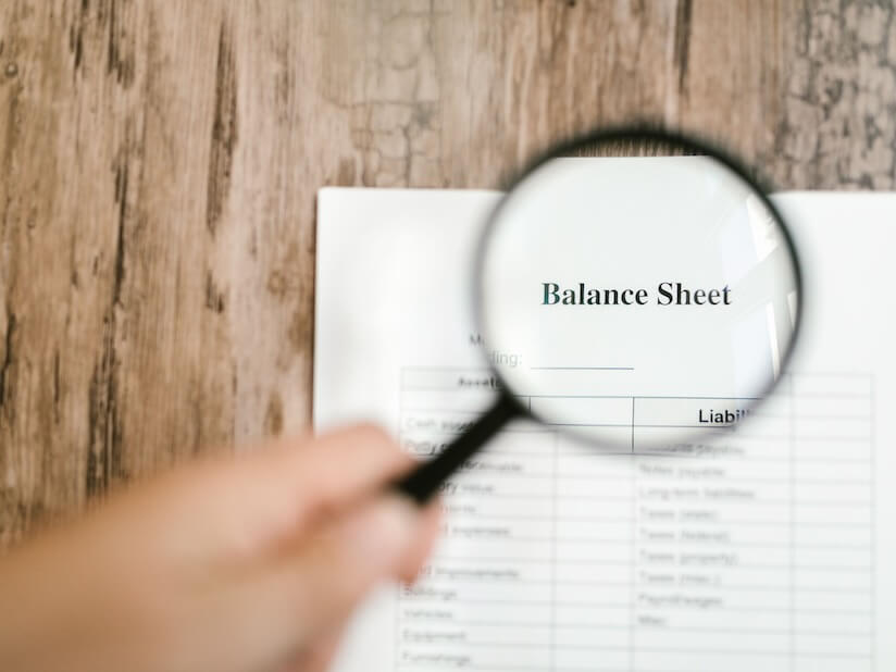 Close-up of a balance sheet document on wooden surface with a magnifying glass held by a hand.