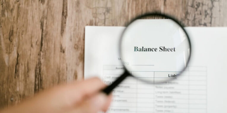 Close-up of a balance sheet document on wooden surface with a magnifying glass held by a hand FEATURED IMAGE