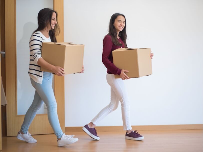 Two young women carrying cardboard boxes indoors, illustrating teamwork and moving in.