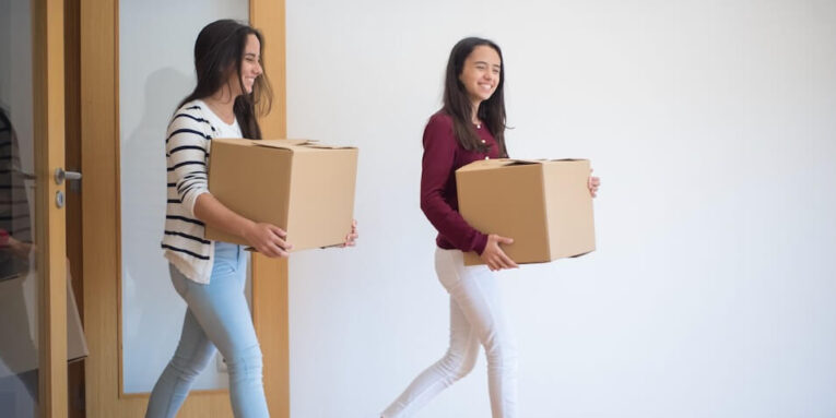 Two young women carrying cardboard boxes indoors, illustrating teamwork and moving in FEATURED IMAGE