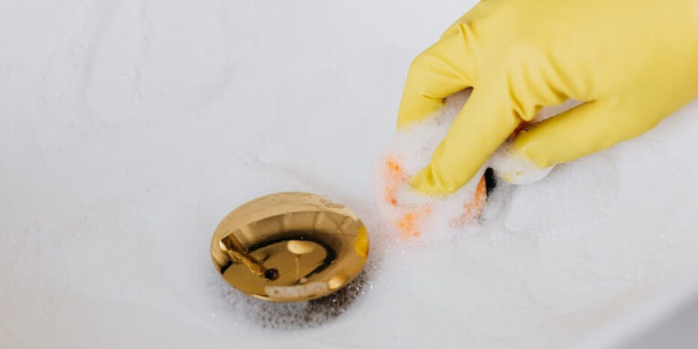 Close-up of a hand in a glove cleaning a foam-covered gold drain in a sink FEATURED IMAGE