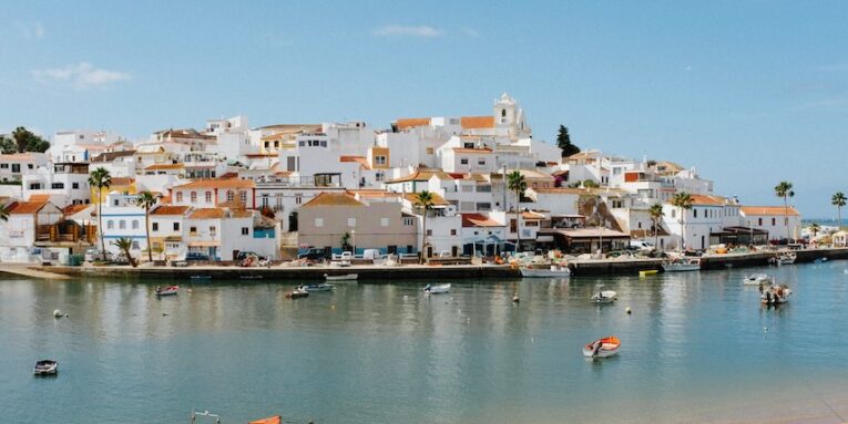 a beach with boats in the water and houses on a hill in the background FEATURED IMAGE