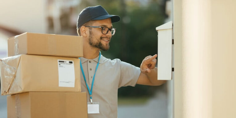 young happy courier checking name on customers mailbox while delivering packages at residential district FEATURED IMAGE