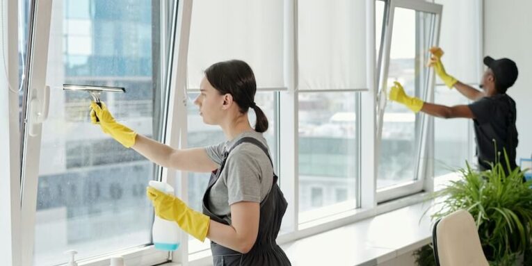 young female cleaner in workwear washing large windows of openspace office against her african american male colleague FEATURED IMAGE