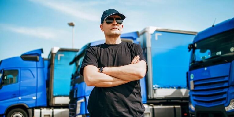 professional truck driver with hat and sunglasses confidently standing in front of big and modern truck bright sunny day people and transportation concept FEATURED IMAGE