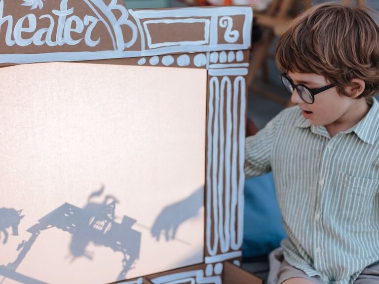 A young boy with glasses playing with a shadow puppet theater indoors.