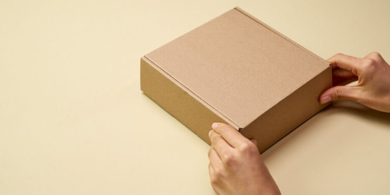 A close-up of hands opening a plain brown cardboard box on a beige surface, perfect for themes of shipping or packaging FEATURED IMAGE