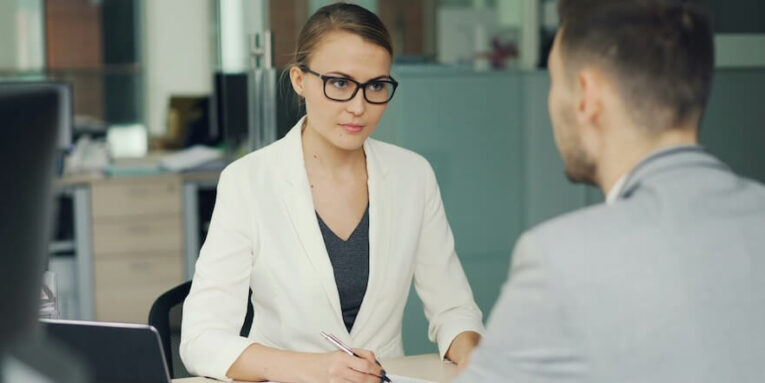 woman in glasses interviews man at office desk FEATURED IMAGE