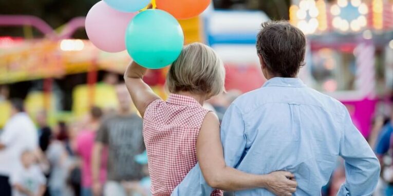senior couple having a good time at the fun fair woman holding colorful balloons sunny summer day rear view FEATURED IMAGE