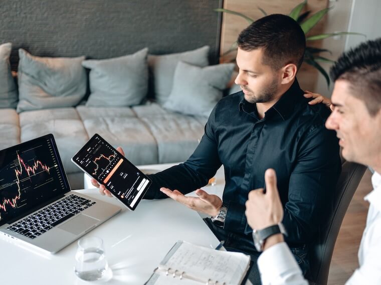 Two businessmen discuss digital trading insights with laptop and tablet in a modern office setting.