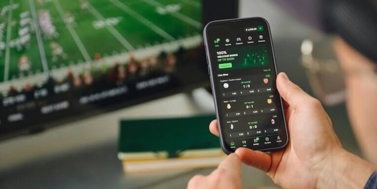 man using phone with sports app near television screen FEATURED IMAGE