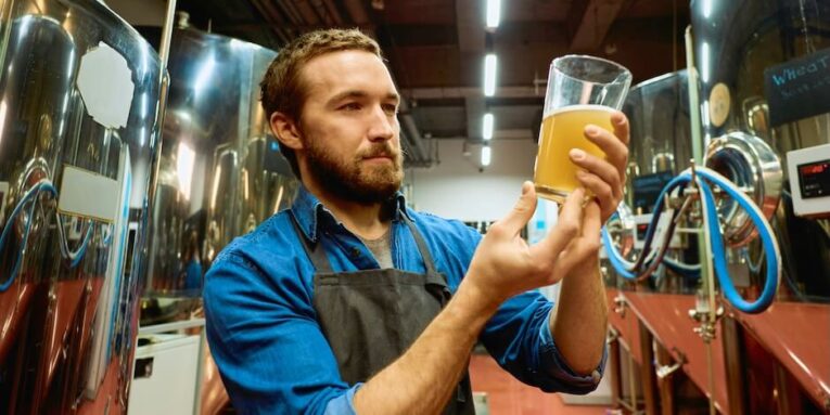 young bearded brewer with glass of beer evaluating its visual characteristics after preparation while standing in front of camera FEATURED IMAGE