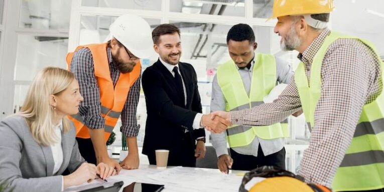 happy construction team shaking hands during conference at office with blueprints FEATURED IMAGE