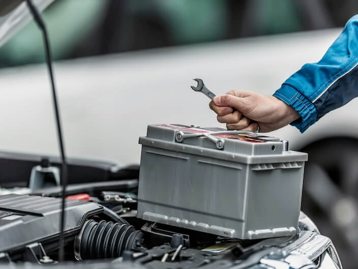 mechanic holds wrench over car battery with engine hood of the car open